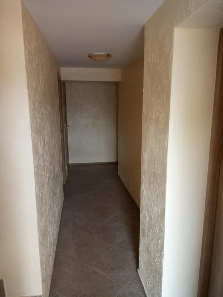 Korytarz z beżowymi ścianami o fakturze trawertynu i ciemnobrązową podłogą z płytek ułożonych w karo, oświetlony przez lampę sufitową.