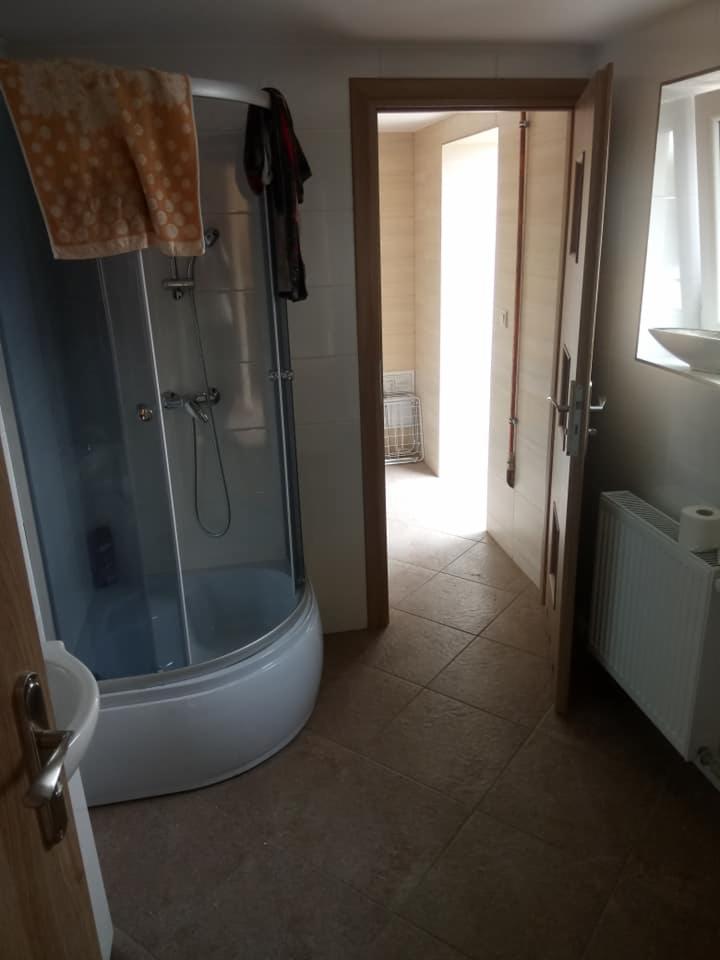 Łazienka z kabiną prysznicową z brązowym ręcznikiem na górze, brązowymi płytkami na podłodze, białym grzejnikiem i otwartymi drzwiami do kolejnej łazienki, widok z perspektywy wejścia.