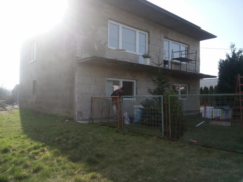 Dom w trakcie ocieplania elewacji, widoczny pracownik przy oknie, surowa ściana z betonu komórkowego, metalowe rusztowanie na balkonie, ogrodzenie z siatki na pierwszym planie.