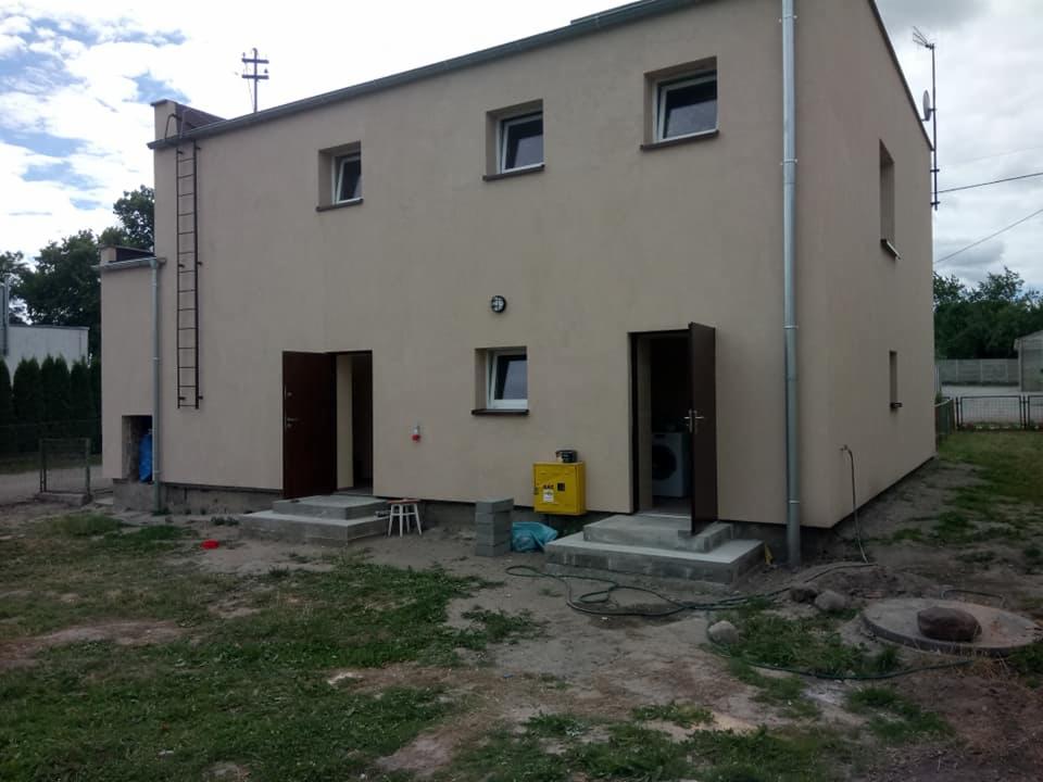 Dwukondygnacyjny budynek o beżowej elewacji z dwiema otwartymi, ciemnobrązowymi drzwiami wejściowymi, zewnętrznymi schodami betonowymi, drabina stalowa na ścianie, żółta skrzynka gazowa i widoczne...