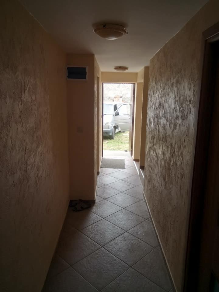 Korytarz z kremowymi ścianami o fakturze i szarą, kwadratową podłogą z płytek, widok na zewnątrz przez otwarte drzwi z zaparkowanym białym vanem.