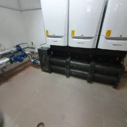 Instalacja trzech białych pomp ciepła De Dietrich z czarnymi rurami w kotłowni w Pszczynie, widoczne rury z izolacją i niebieskimi zaworami.