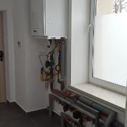 Nowoczesny, wiszący piec gazowy marki Vaillant z rozbudowaną instalacją rur i zaworów, umieszczony w jasnym pomieszczeniu z szarą podłogą i oknem z częściowo matową szybą.