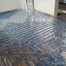 Niebieskie rury ogrzewania podłogowego ułożone na folii refleksyjnej z siatką, widoczne fragmenty ścian w surowym stanie.