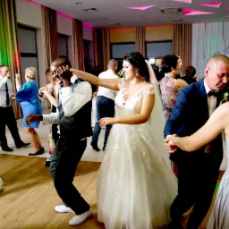 Radosna zabawa weselna: uśmiechnięta panna młoda w koronkowej sukni tańczy z gośćmi na parkiecie z drewnianą podłogą, oświetlonym kolorowymi światłami.