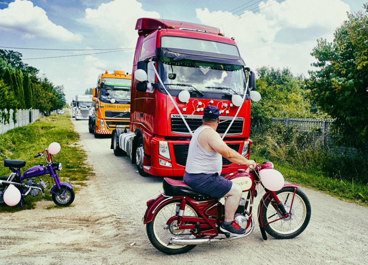 Udekorowana ciężarówka Volvo z białymi wstążkami i sercami, jadąca w konwoju, poprzedzana przez mężczyznę na czerwonym motocyklu WSK i fioletowy motorower Romet z balonami.