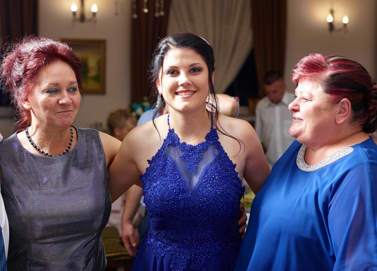 Uśmiechnięta młoda kobieta w kobaltowej sukni z koronkową górą, pozująca z dwiema starszymi kobietami w pomieszczeniu z brązowymi zasłonami i oświetleniem.