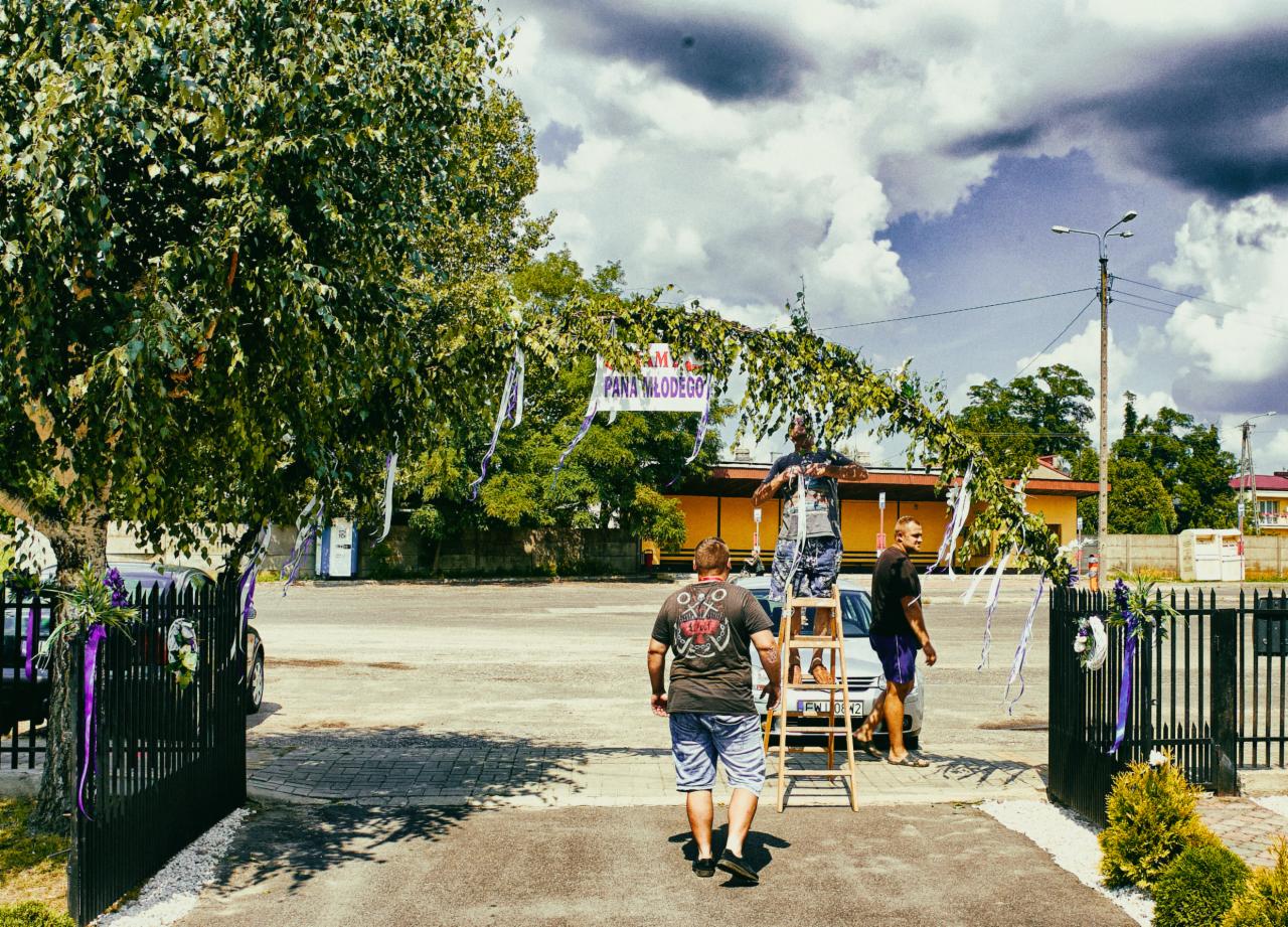 Mężczyzna na drabinie dekoruje wejście zielonymi gałązkami i białymi wstążkami, baner z napisem 'Dla Młodej Pary', przygotowania do uroczystości.