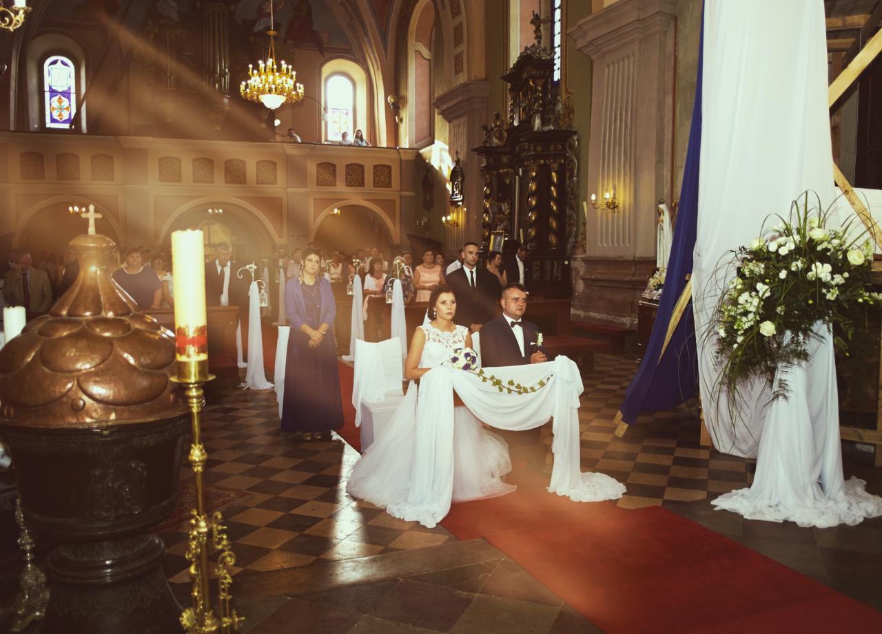 Para młoda siedzi w kościele podczas ceremonii ślubnej, oświetlona promieniami słońca wpadającymi przez okno, widoczny tłum gości i dekoracje kwiatowe.