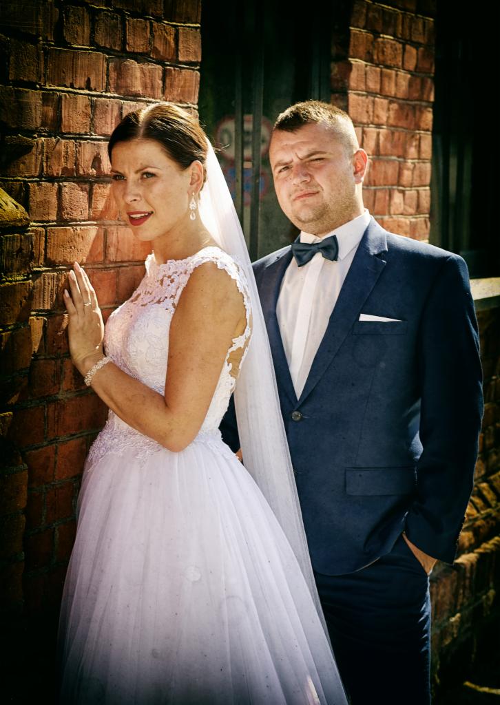 Panna młoda w koronkowej sukni ślubnej z welonem, oparta o ceglaną ścianę, obok pan młody w granatowym garniturze z muchą, pozujący na tle muru z oknem.