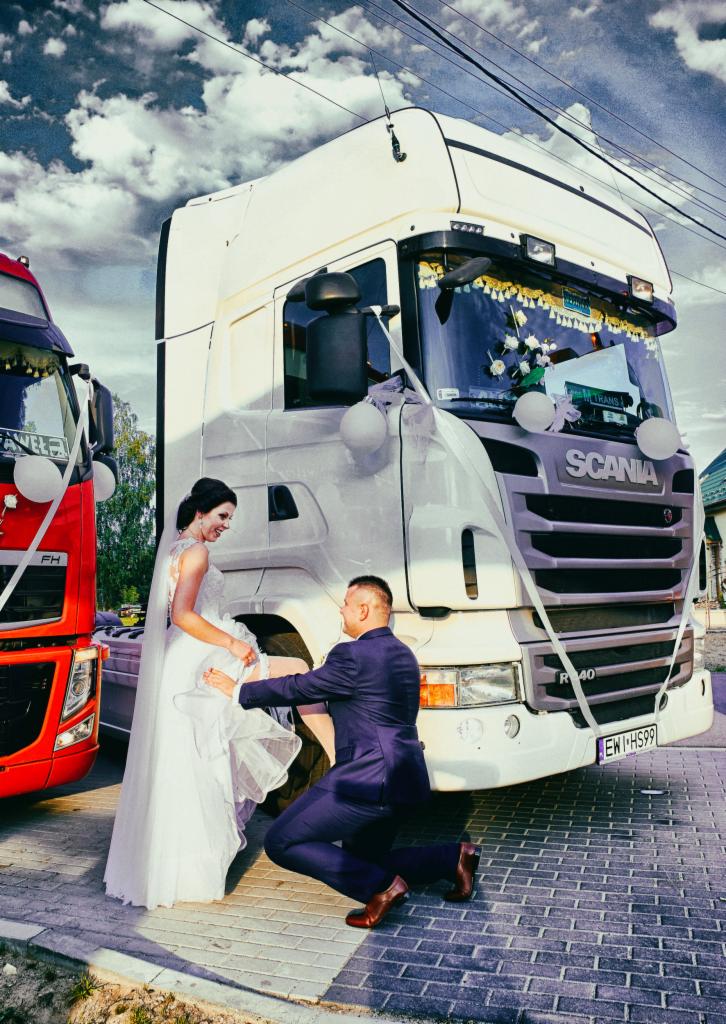 Para młoda pozuje na tle udekorowanej ciężarówki Scania R40 z białymi balonami i kwiatami, oblubieniec klęczy zakładając podwiązkę oblubienicy, w tle druga ciężarówka FH