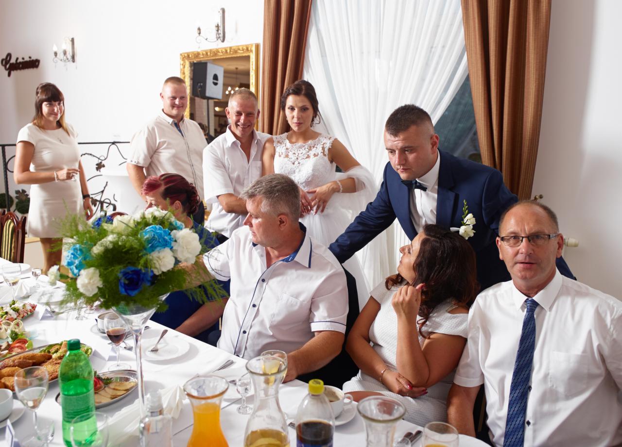 Panna młoda w białej sukni pozuje z gośćmi weselnymi przy stole z przekąskami i bukietem niebiesko-białych kwiatów.