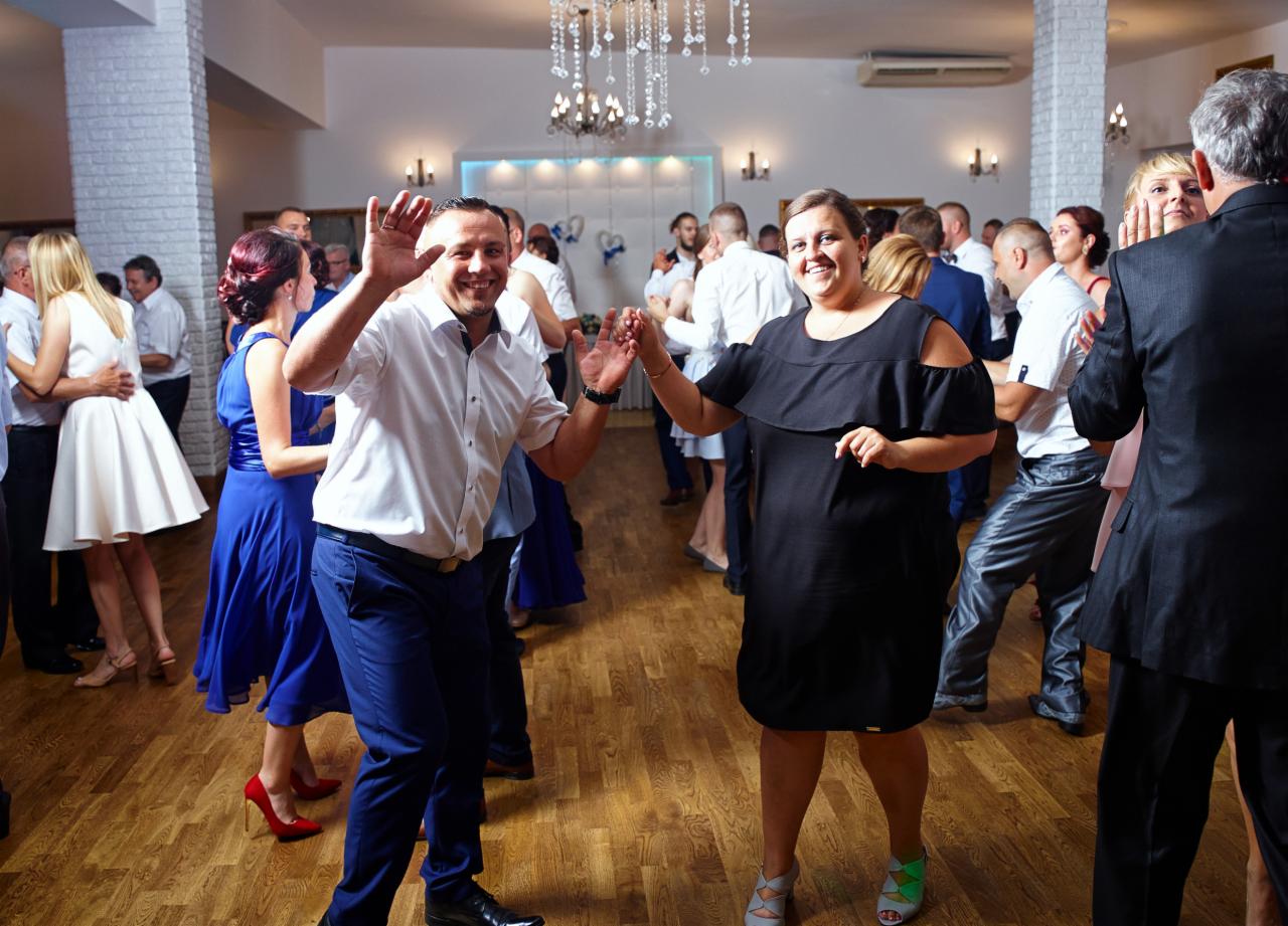 Rozbawieni goście weselni tańczą na parkiecie, mężczyzna w białej koszuli i niebieskich spodniach macha ręką, kobieta w czarnej sukience uśmiecha się, w tle bawią się inne pary.