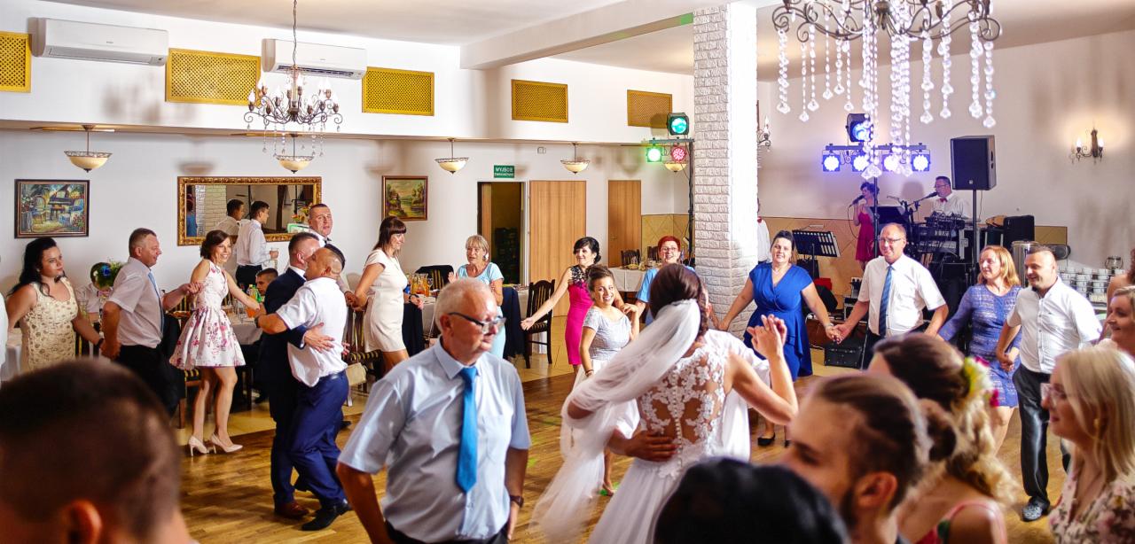 Roztańczony tłum gości weselnych w sali bankietowej, w centrum panna młoda w białej sukni z koronkowymi plecami, w tle zespół muzyczny i dekoracje świetlne.