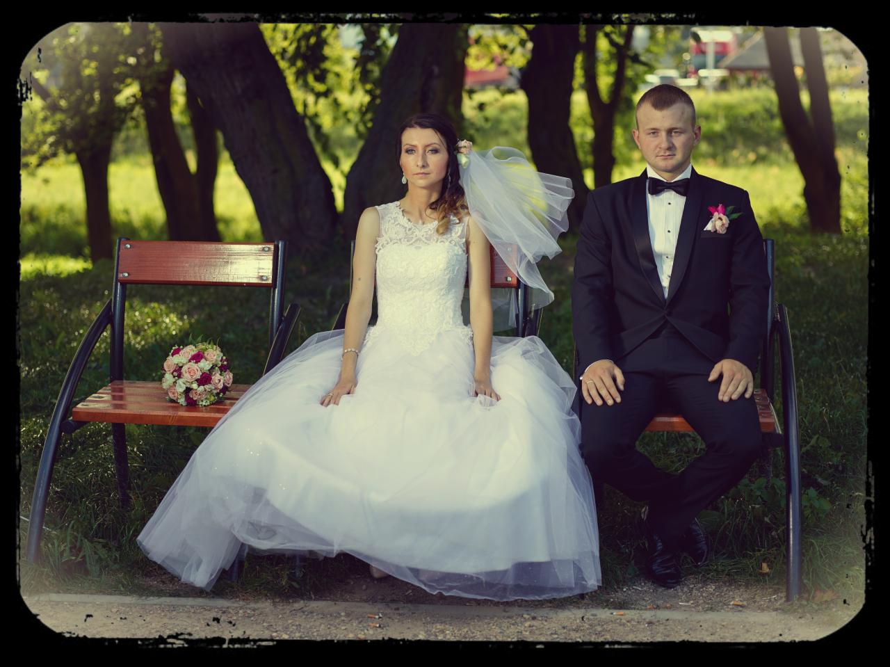 Para młoda siedząca na ławce w parku: panna młoda w białej sukni z koronkową górą i welonem, obok pan młody w czarnym smokingu z muchą i butonierką, bukiet ślubny na ławce.