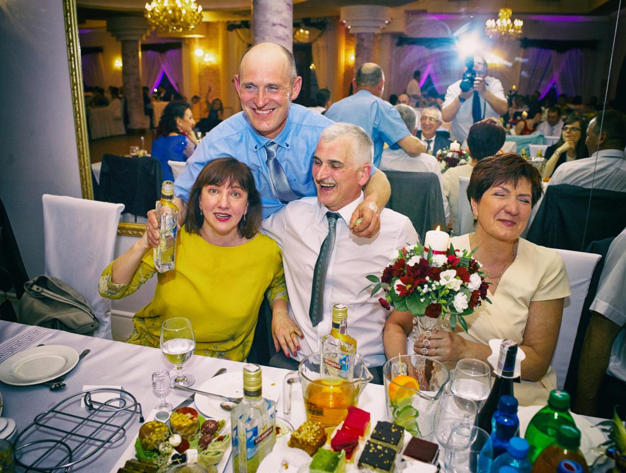 Grupa gości weselnych uśmiechnięta przy stole z przekąskami i napojami, w tle fotograf z lampą błyskową.