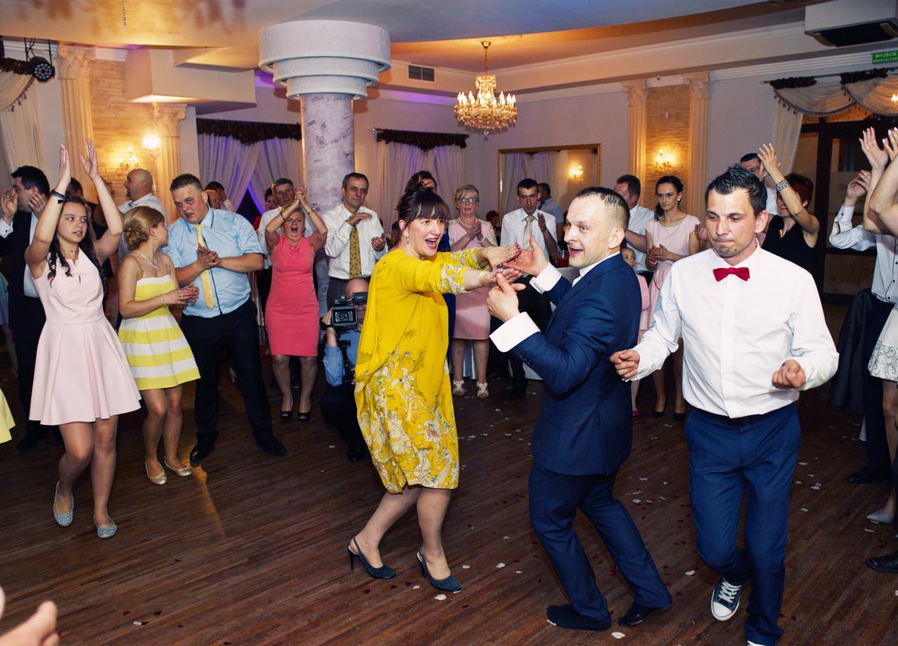 Para w żółtej sukience i granatowym garniturze tańczy podczas wesela, otoczona przez gości z podniesionymi rękami, w tle kolumny i kryształowy żyrandol