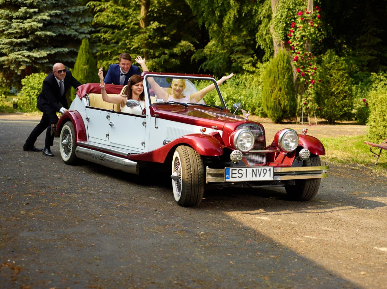 Para młoda w czerwono-białym kabriolecie z lat 30. XX wieku, z otwartymi ramionami, eskortowana przez mężczyznę w garniturze, na tle zieleni.