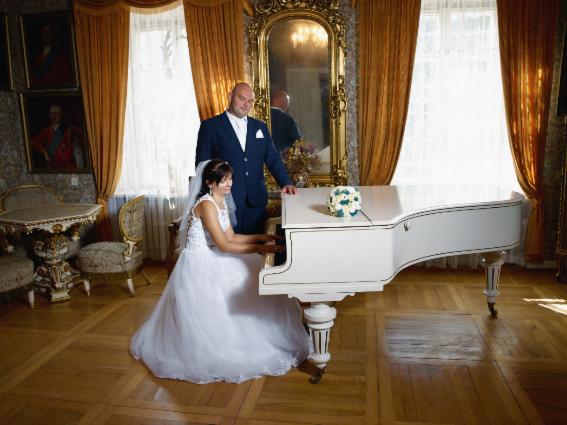 Panna młoda w białej sukni gra na białym fortepianie w eleganckim wnętrzu, obok stoi pan młody w granatowym garniturze, w tle złote zasłony i lustro.
