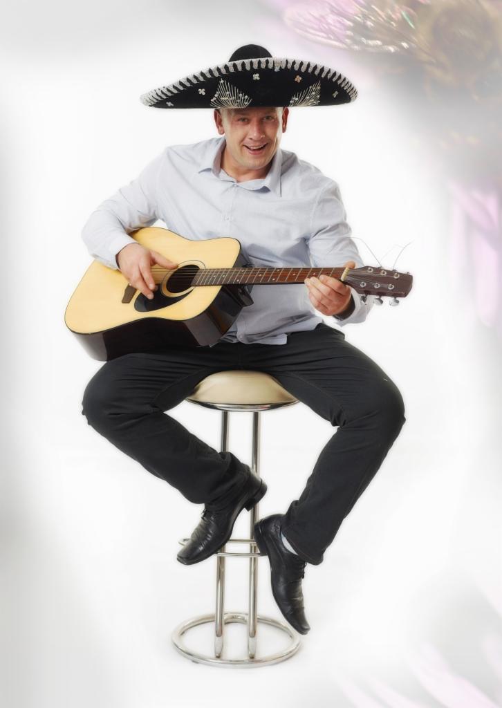 Uśmiechnięty mężczyzna w sombrero siedzi na stołku i gra na gitarze akustycznej, ubrany w jasną koszulę i ciemne spodnie.