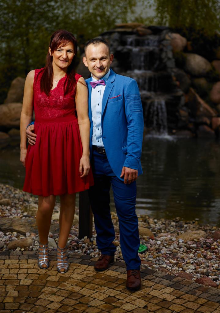Para pozuje do zdjęcia na tle wodospadu w parku, kobieta w czerwonej sukience i srebrnych butach, mężczyzna w niebieskim garniturze i bordowej muszce.
