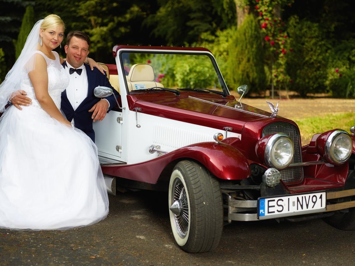 Para młoda pozuje obok bordowo-białego samochodu retro z ozdobną figurką orła na masce, w tle zieleń ogrodu.