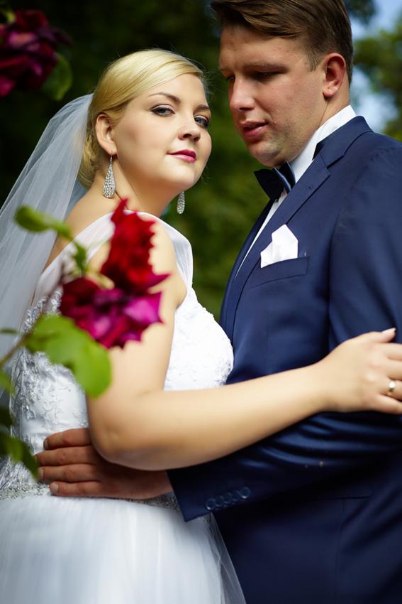Para młoda w objęciach, panna młoda patrzy w obiektyw, w tle zielona roślinność i rozkwitające ciemnoczerwone róże.