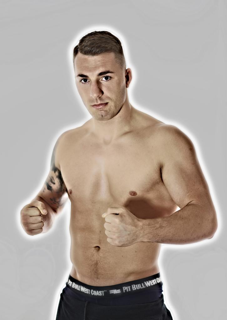 Portret muskularnego mężczyzny bez koszuli, z tatuażem na ramieniu, w bieliźnie sportowej marki Pit Bull West Coast, na jasnym tle.