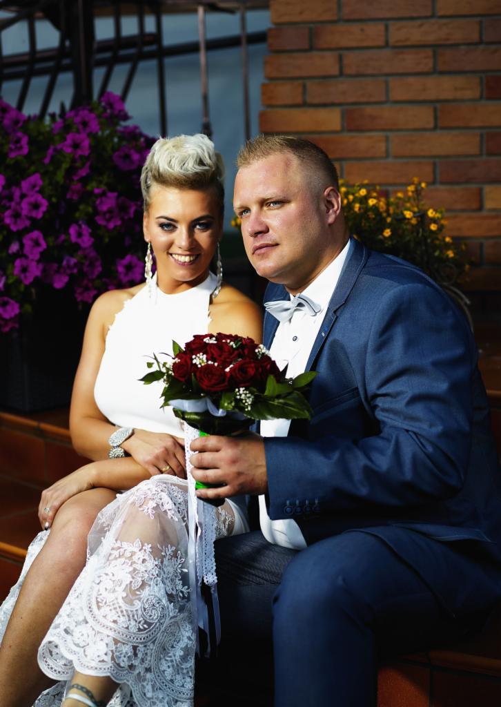 Para młoda siedzi na schodach przed ceglaną ścianą. Panna młoda w białej sukni z koronkowym dołem i bukietem czerwonych róż, pan młody w niebieskim garniturze z muchą.