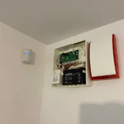 Otwarta centrala alarmowa na ścianie z widoczną płytką drukowaną, akumulatorem Alarmtec i czujnikiem ruchu obok.