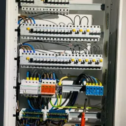 Otwarta rozdzielnica elektryczna z zainstalowanymi bezpiecznikami, zabezpieczeniami przepięciowymi i listwami zaciskowymi, widoczne narzędzia: nóż monterski i szczypce.