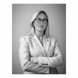 Portret kobiety w okularach i jasnym garniturze, z założonymi rękami, na neutralnym tle, czarno-biały.