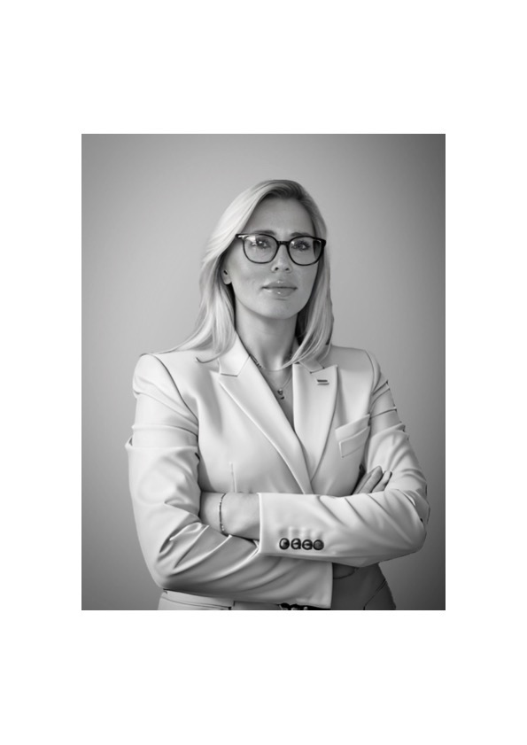 Portret kobiety w okularach i jasnym garniturze, z założonymi rękami, na neutralnym tle, czarno-biały.