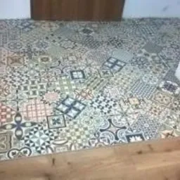 Wzorzysta, patchworkowa podłoga z płytek ceramicznych w odcieniach szarości, błękitu i beżu, ułożona na drewnianym podłożu przy wejściu do pomieszczenia, z widocznym fragmentem drzwi i butem...