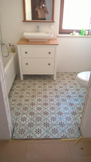 Ułożone, wzorzyste płytki podłogowe w łazience, widoczna biała szafka z umywalką i lustro z odbiciem osoby fotografującej.