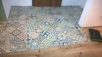 Wzorzysta, patchworkowa podłoga z płytek ceramicznych w odcieniach szarości, błękitu i beżu, ułożona na drewnianym podłożu przy wejściu do pomieszczenia, z widocznym fragmentem drzwi i butem...