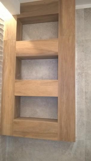 Wnęka z drewnianym obramowaniem, wyłożona szarą płytką, wkomponowana w ścianę pokrytą płytkami w podobnym odcieniu.