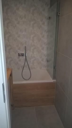 Wykończona łazienka z wanną obudowaną drewnopodobnymi panelami, przeszklonym parawanem nawannowym i geometrycznymi płytkami na ścianie.