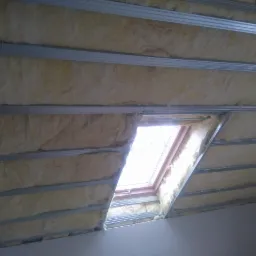 Poddasze z oknem dachowym w trakcie ocieplania wełną mineralną, widoczne metalowe profile konstrukcyjne i fragment żarówki.