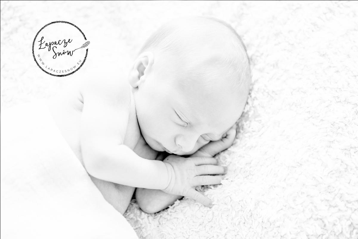 Czarno-białe zdjęcie noworodka śpiącego na puszystym, białym kocyku z delikatnie zaciśniętą dłonią i widocznym logo fotografa w lewym górnym rogu kadru.