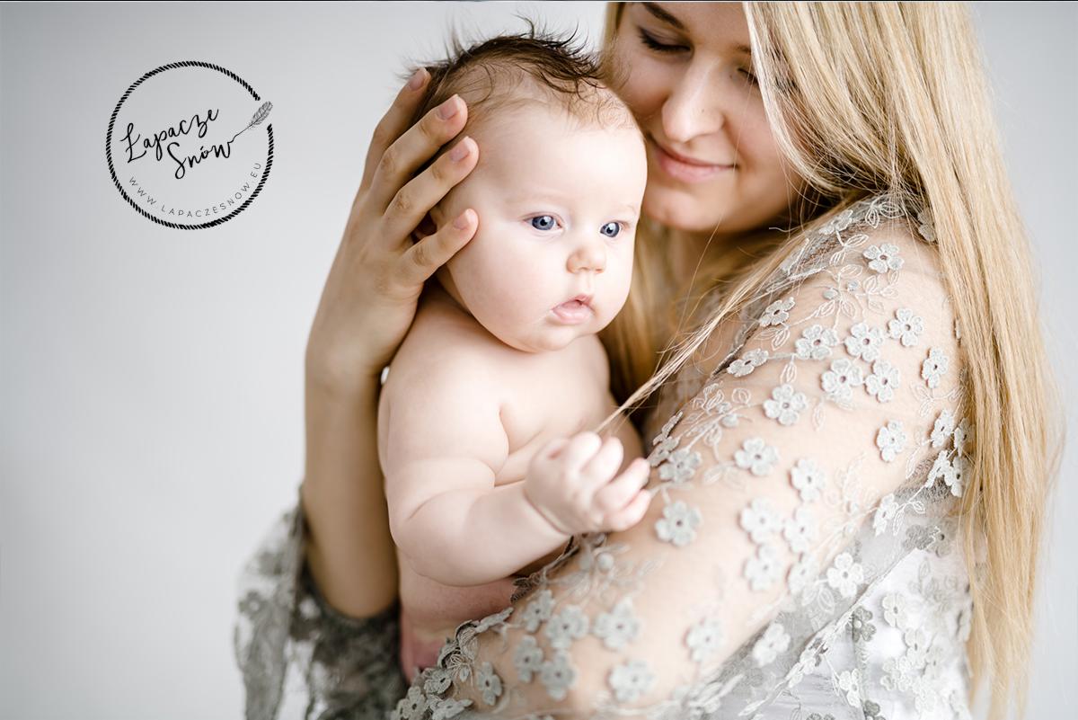 Portret matki trzymającej niemowlę na rękach, delikatne oświetlenie, kobieta w bluzce z kwiatowym wzorem, logo fotografa w lewym górnym rogu.