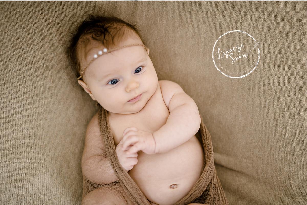 Niemowlę z niebieskimi oczami, ubrane w brązową chustę i opaskę z perełkami, leżące na beżowym tle z widocznym logo fotografa.