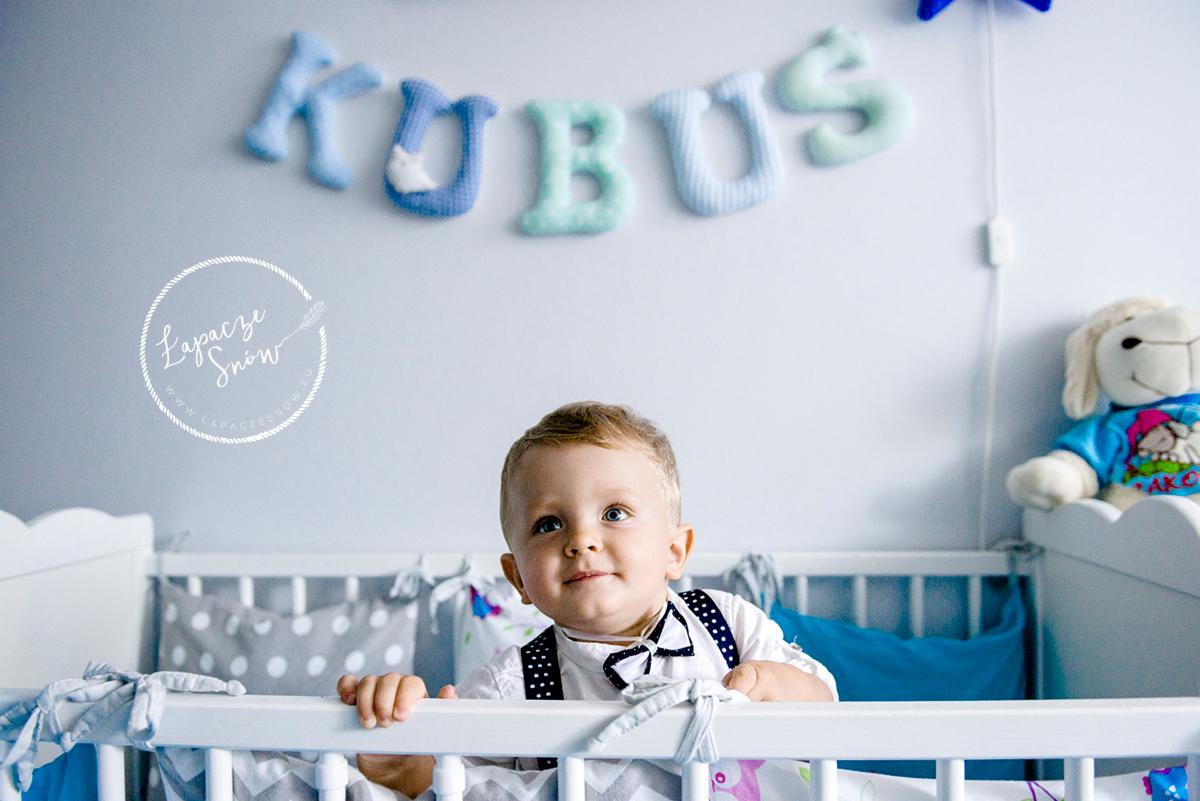 Uśmiechnięty chłopiec w muszce i szelkach stoi w białym łóżeczku dziecięcym, za nim na ścianie wiszą dekoracyjne litery układające się w imię 'KUBUS', a w tle widoczny pluszowy baranek.