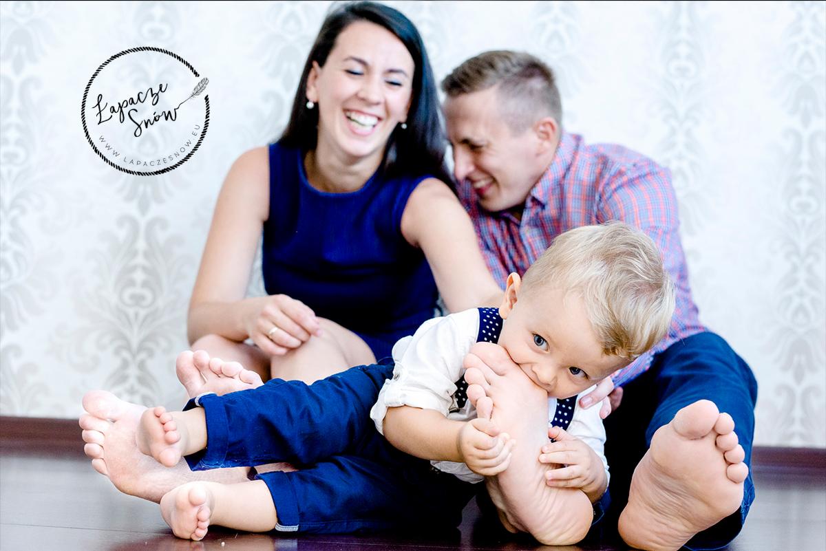 Radosna rodzina: mama i tata siedzą na podłodze, śmiejąc się, podczas gdy ich mały syn z blond włosami gryzie stopę taty, na tle ściany z delikatnym wzorem i logo fotografa.