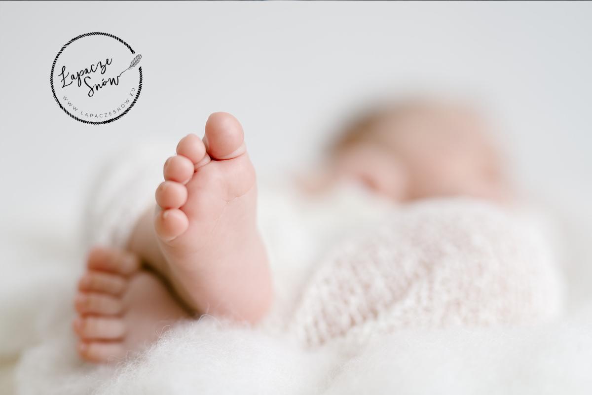 Małe stópki noworodka na białym, puszystym kocyku z widocznym logo fotografa w lewym górnym rogu.