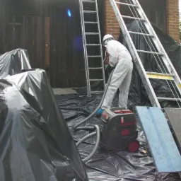 Pracownik w kombinezonie ochronnym, z maską i kasku, odkurza powierzchnię budynku zabezpieczoną folią, używając przemysłowego odkurzacza. W tle widoczne dwie drabiny opierające się o budynek...