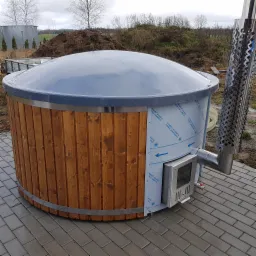 Drewniana beczka z piecem zewnętrznym i kominem na posesji z kostki brukowej, zabezpieczona folią ochronną, w tle pole i turbiny wiatrowe.