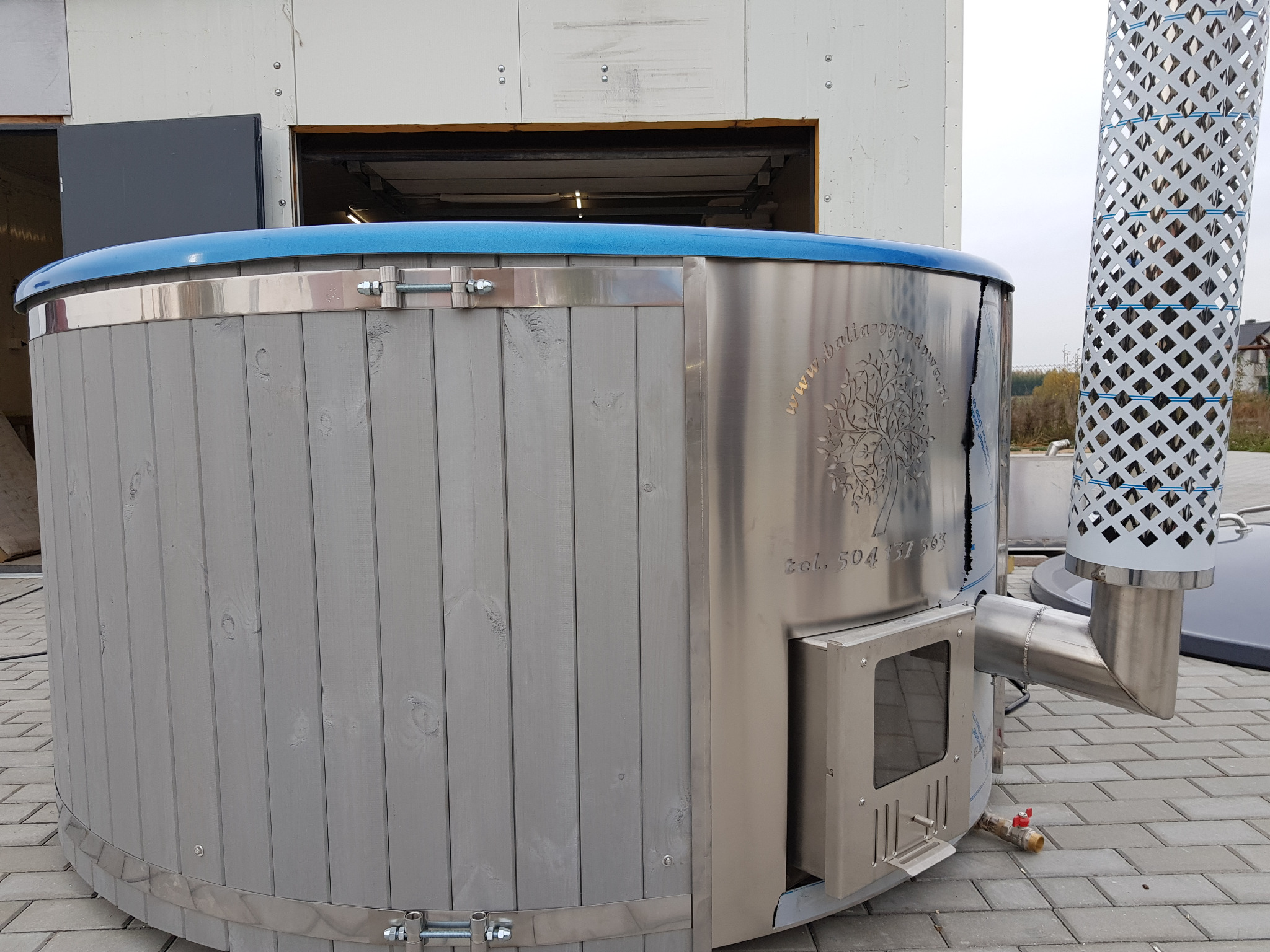 Drewniana balia ogrodowa z piecem na drewno, obudowana metalem, z kominem zabezpieczonym ażurową osłoną.