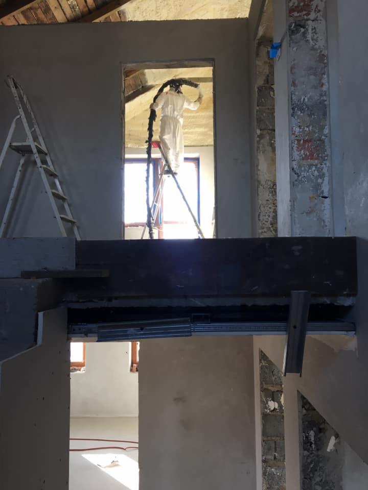 Pracownik w kombinezonie ochronnym aplikuje izolację natryskową na poddaszu, stojąc na drabinie; widoczna surowa cegła i fragment konstrukcji dachu.