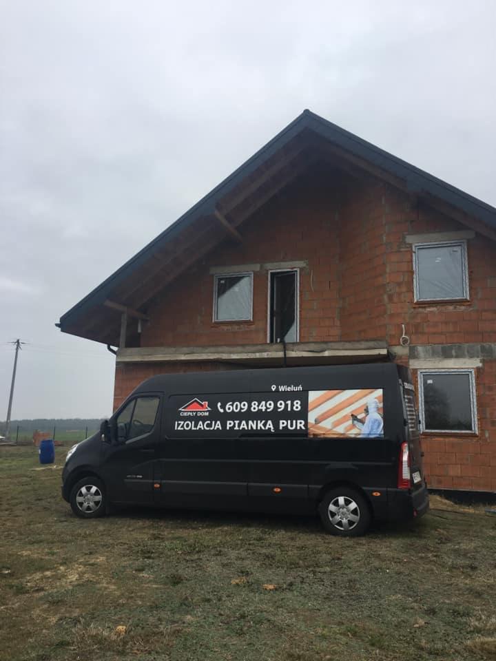 Czarny samochód dostawczy z reklamą firmy ociepleniowej na tle surowego, ceglanego domu w trakcie budowy, widoczne niezabezpieczone okna.